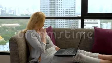 一位优雅的<strong>女士</strong>躺在沙发上，坐在全景窗旁，俯瞰摩天大楼，并使<strong>用</strong>她的<strong>笔记本电脑</strong>